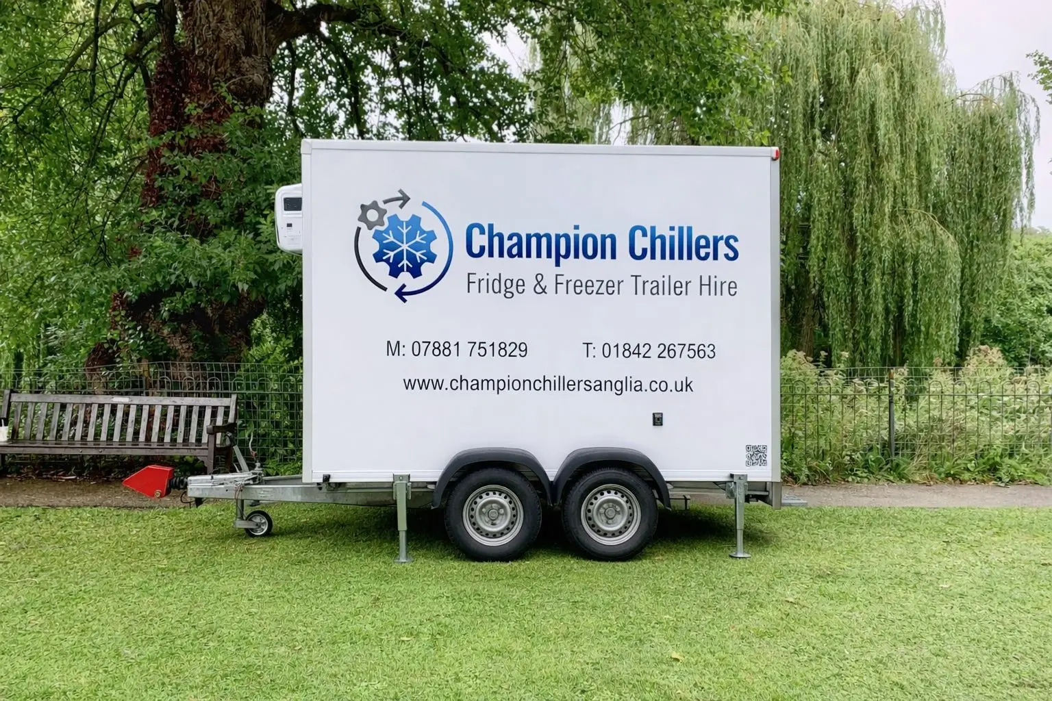 Champion Chillers fridge and freezer trailer hire unit parked on grass beside a park bench and willow trees.