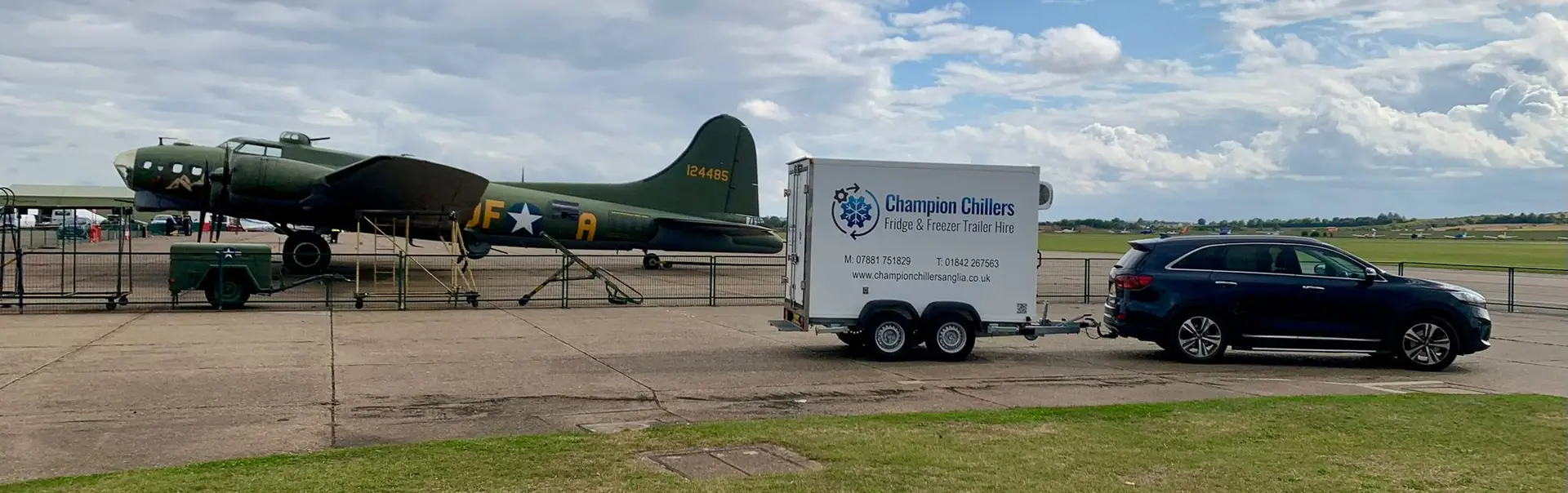 Car towing a Champion Chillers fridge and freezer trailer hire unit past a vintage WWII bomber at an airfield.
