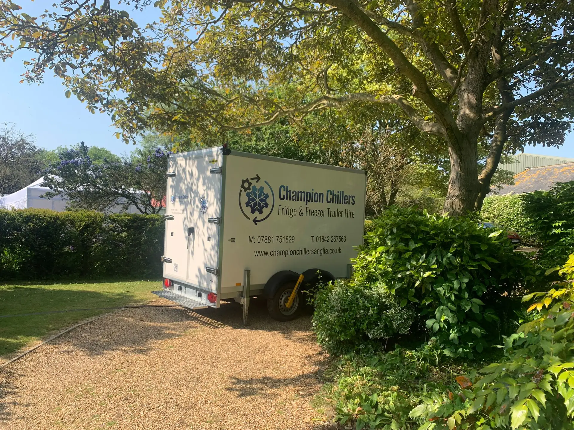 Champion Chillers fridge and freezer trailer hire unit parked on a gravel driveway in a garden, with a marquee behind.