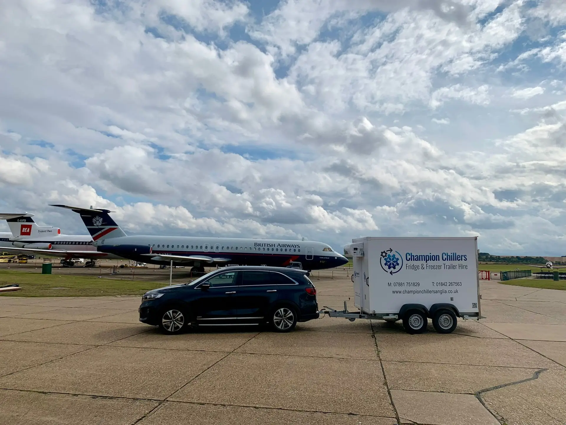Car towing a Champion Chillers fridge and freezer trailer hire unit across an airfield, with vintage aircraft behind.